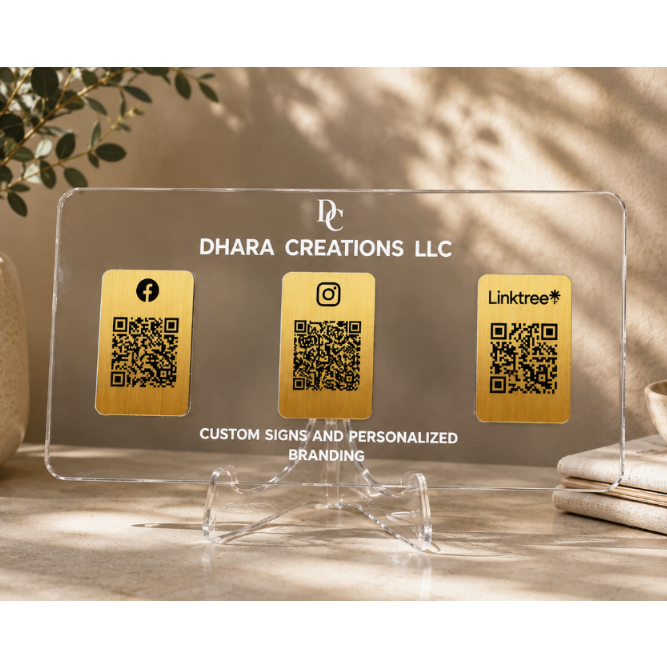 A clear acrylic custom business sign on a clear acrylic stand, featuring three gold QR code panels labeled with Facebook, Instagram, and Linktree, with the text 'DHARA CREATIONS LLC' and 'CUSTOM SIGNS AND PERSONALIZED BRANDING'. The sign is styled on a tabletop with a potted plant and books in the background.