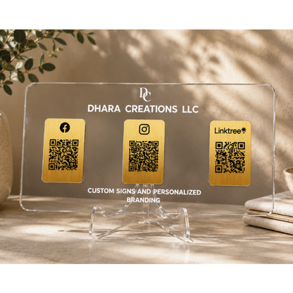 A clear acrylic custom business sign on a clear acrylic stand, featuring three gold QR code panels labeled with Facebook, Instagram, and Linktree, with the text 'DHARA CREATIONS LLC' and 'CUSTOM SIGNS AND PERSONALIZED BRANDING'. The sign is styled on a tabletop with a potted plant and books in the background.