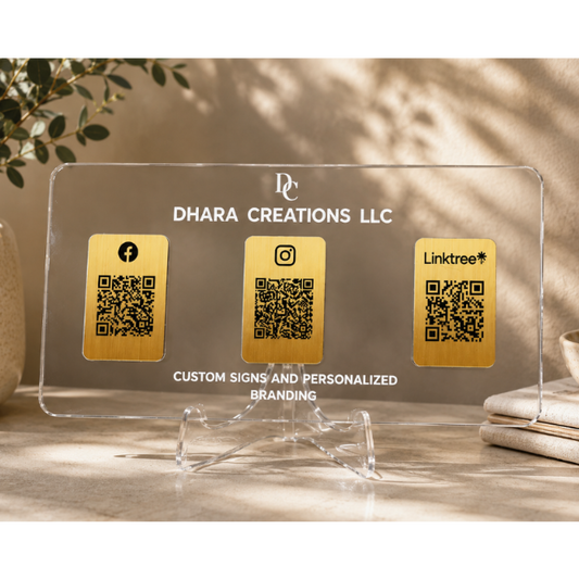 A clear acrylic custom business sign on a clear acrylic stand, featuring three gold QR code panels labeled with Facebook, Instagram, and Linktree, with the text 'DHARA CREATIONS LLC' and 'CUSTOM SIGNS AND PERSONALIZED BRANDING'. The sign is styled on a tabletop with a potted plant and books in the background.